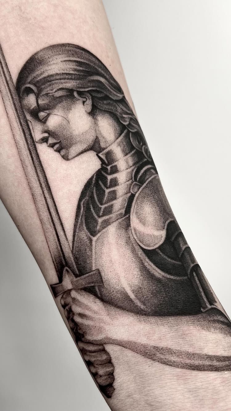 7 Knight Style Tattoo Ideas for 2026 5 A black and grey realism tattoo of a female knight in armor holding a sword on an arm.