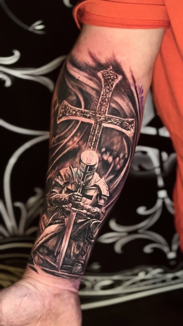 7 Knight Style Tattoo Ideas for 2026 6 A detailed black and grey tattoo of a kneeling knight in front of a large cross.
