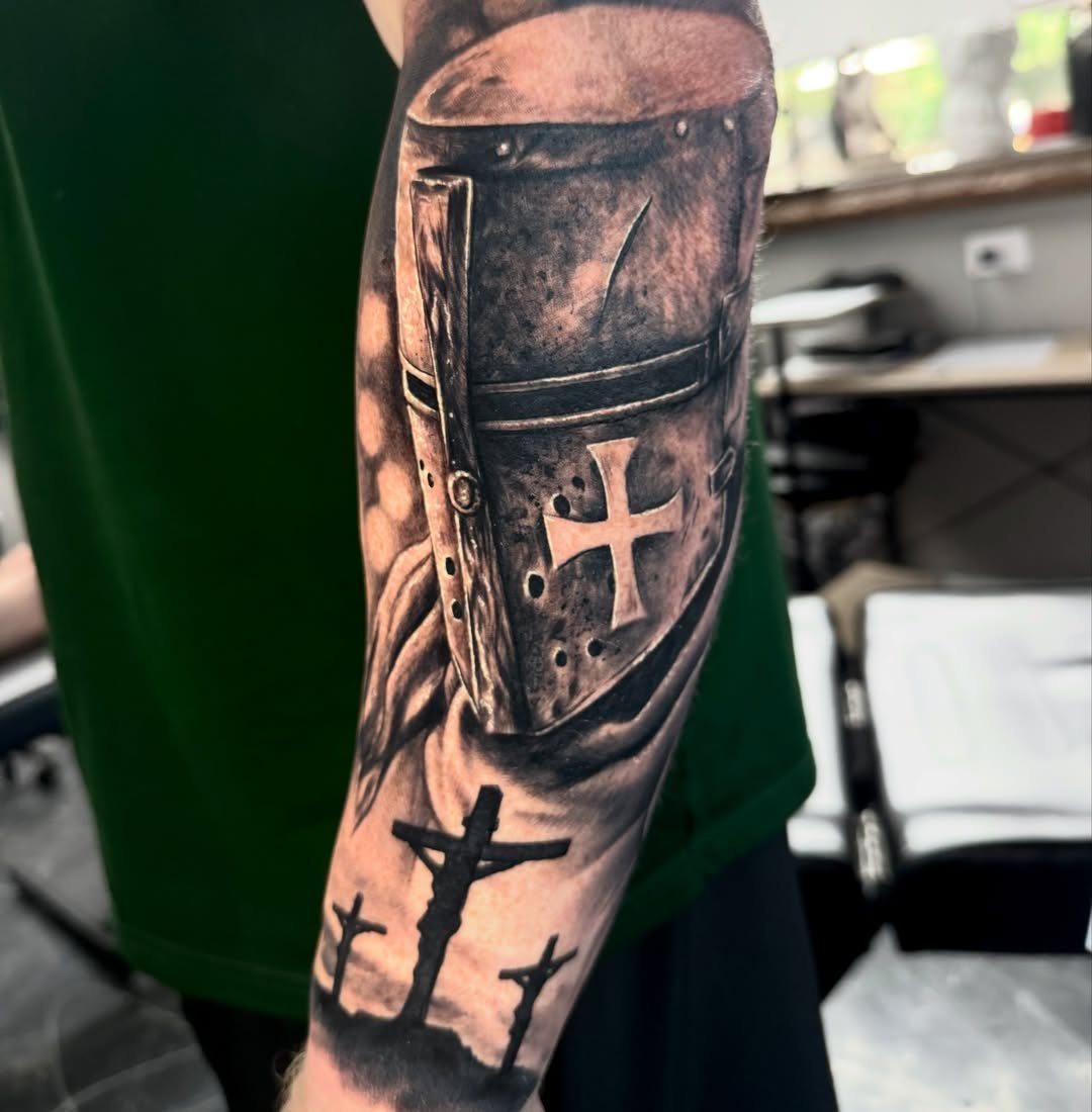 7 Knight Style Tattoo Ideas for 2026 1 A black and grey realism tattoo of a knight helmet and three crosses on an arm.