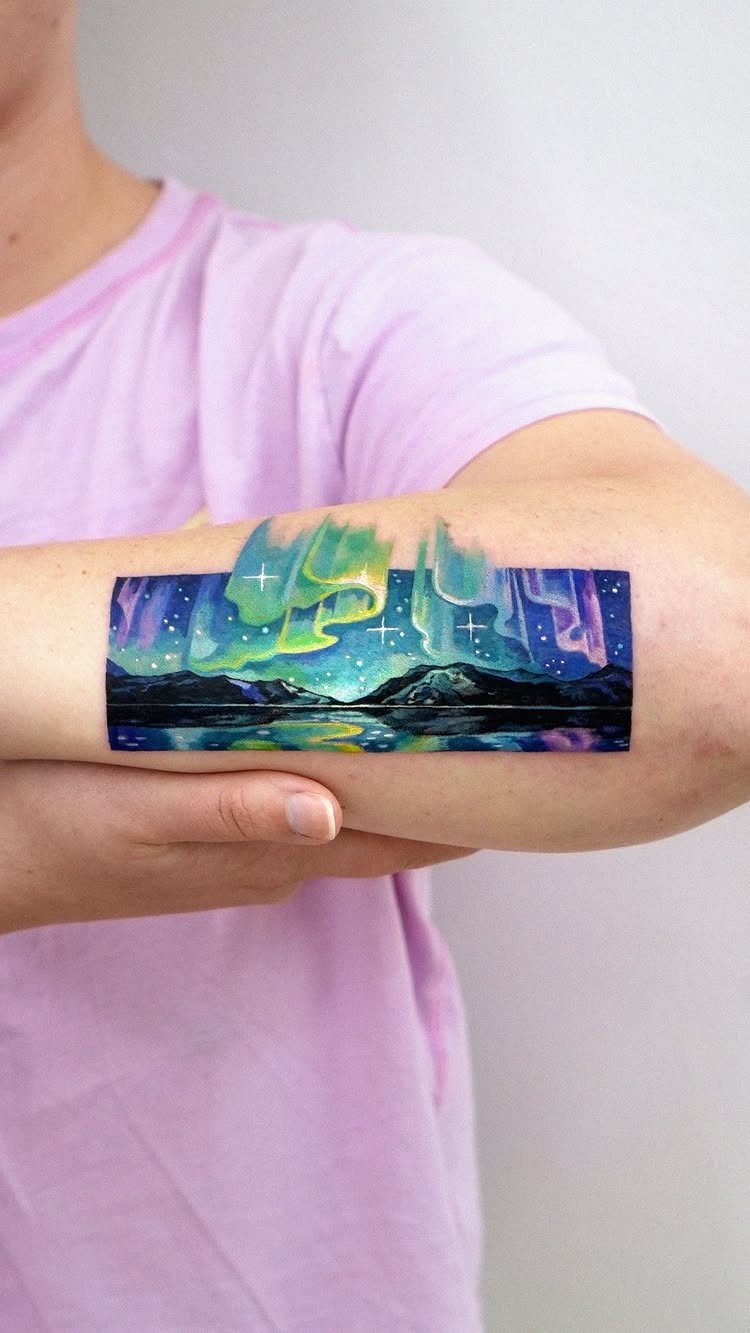 A vibrant aurora borealis landscape tattoo on a person's upper arm with mountains and stars.