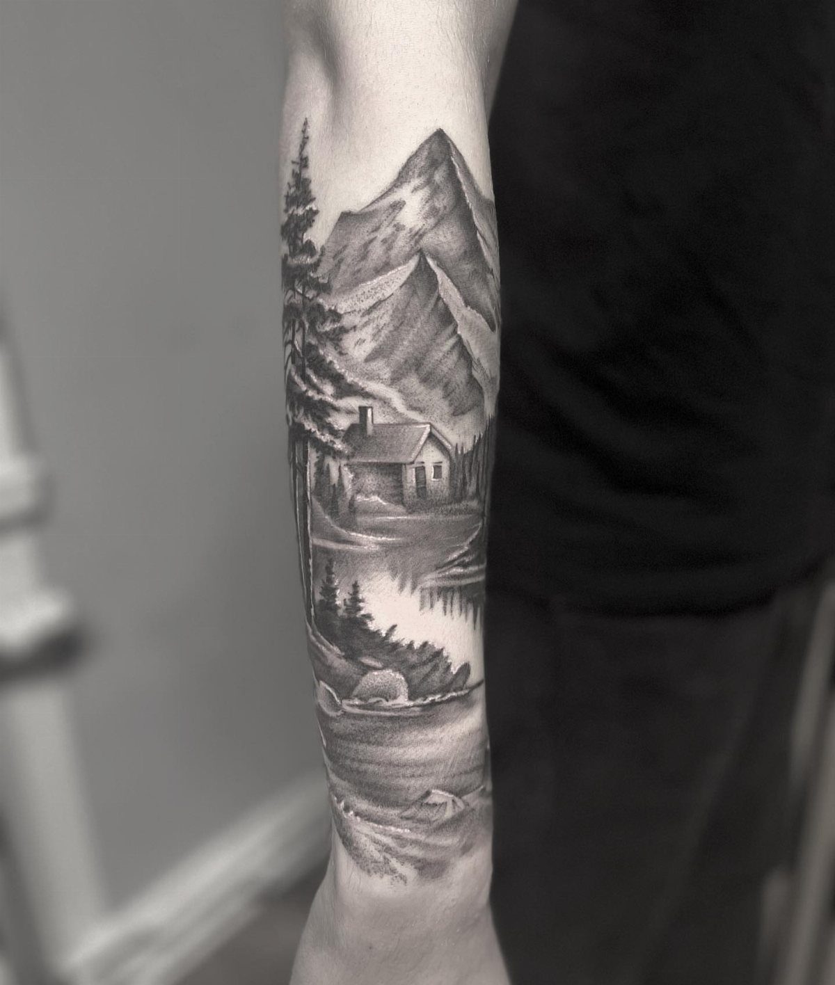 A black and grey landscape tattoo on a forearm featuring mountains, a cabin, and trees.