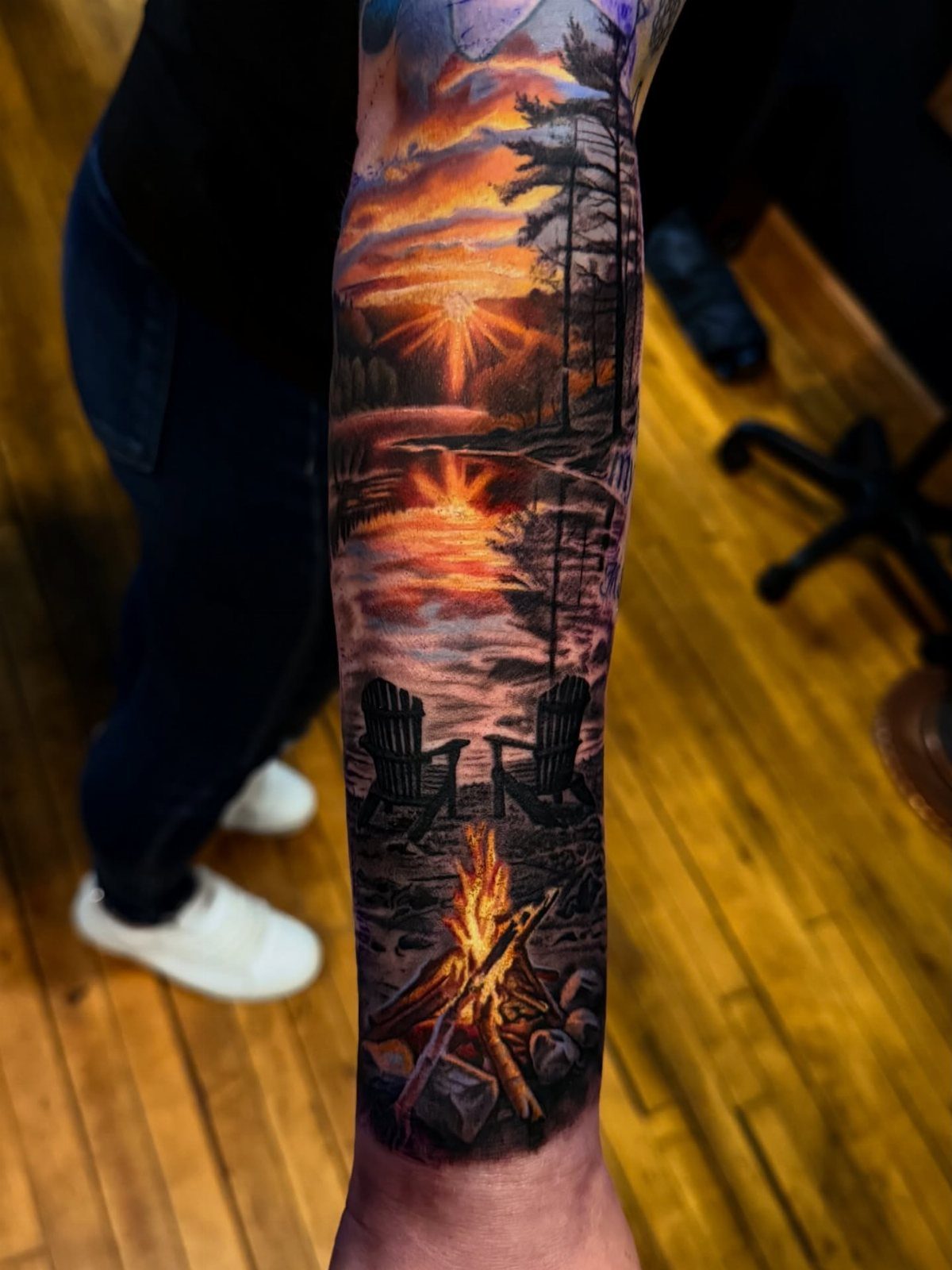 7 Landscape Tattoo Sleeve Ideas for Your Next Ink 6 A detailed arm tattoo sleeve showing a colorful sunset landscape with a campfire and lake.
