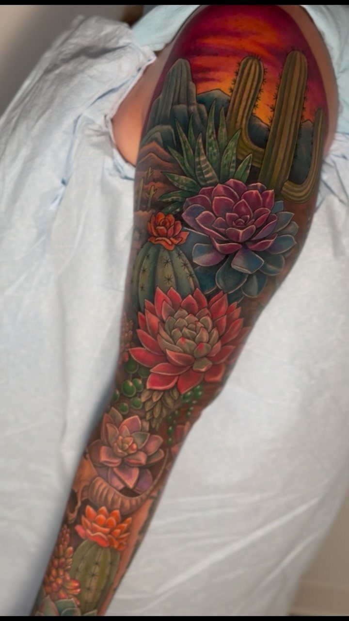 7 Landscape Tattoo Sleeve Ideas for Your Next Ink 2 A colorful tattoo sleeve on an arm depicting various cacti and succulents against a sunset sky.