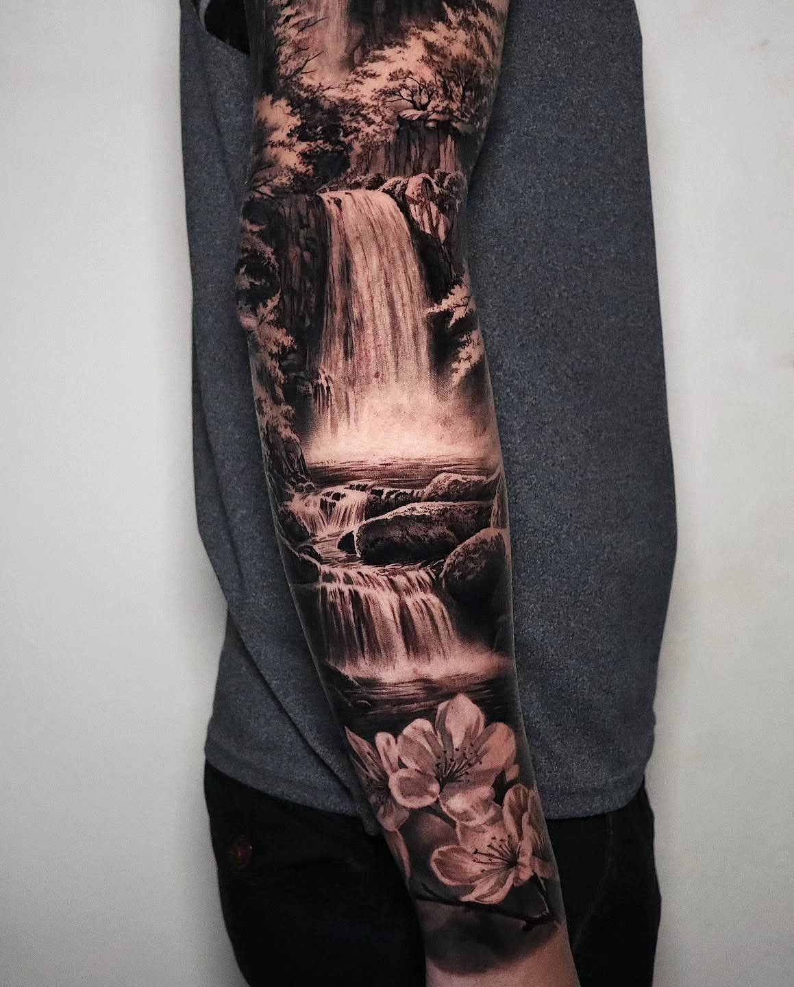 7 Landscape Tattoo Sleeve Ideas for Your Next Ink 4 A detailed black and grey tattoo sleeve depicting a waterfall landscape with trees and flowers.