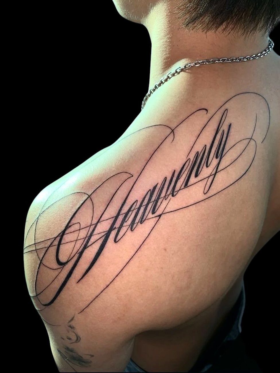 Large Back Script Tattoo: 5 Styles That Fill the Space 2 Large black cursive script tattoo reading Heavenly on a person's upper back and shoulder.