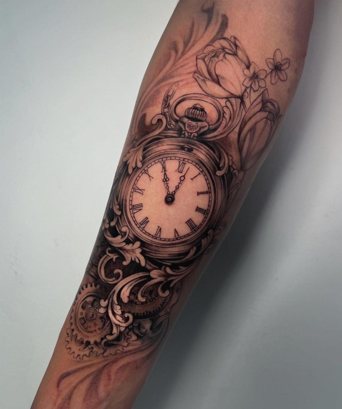 A detailed black and grey tattoo of an ornate pocket watch with filigree and flowers on an arm.