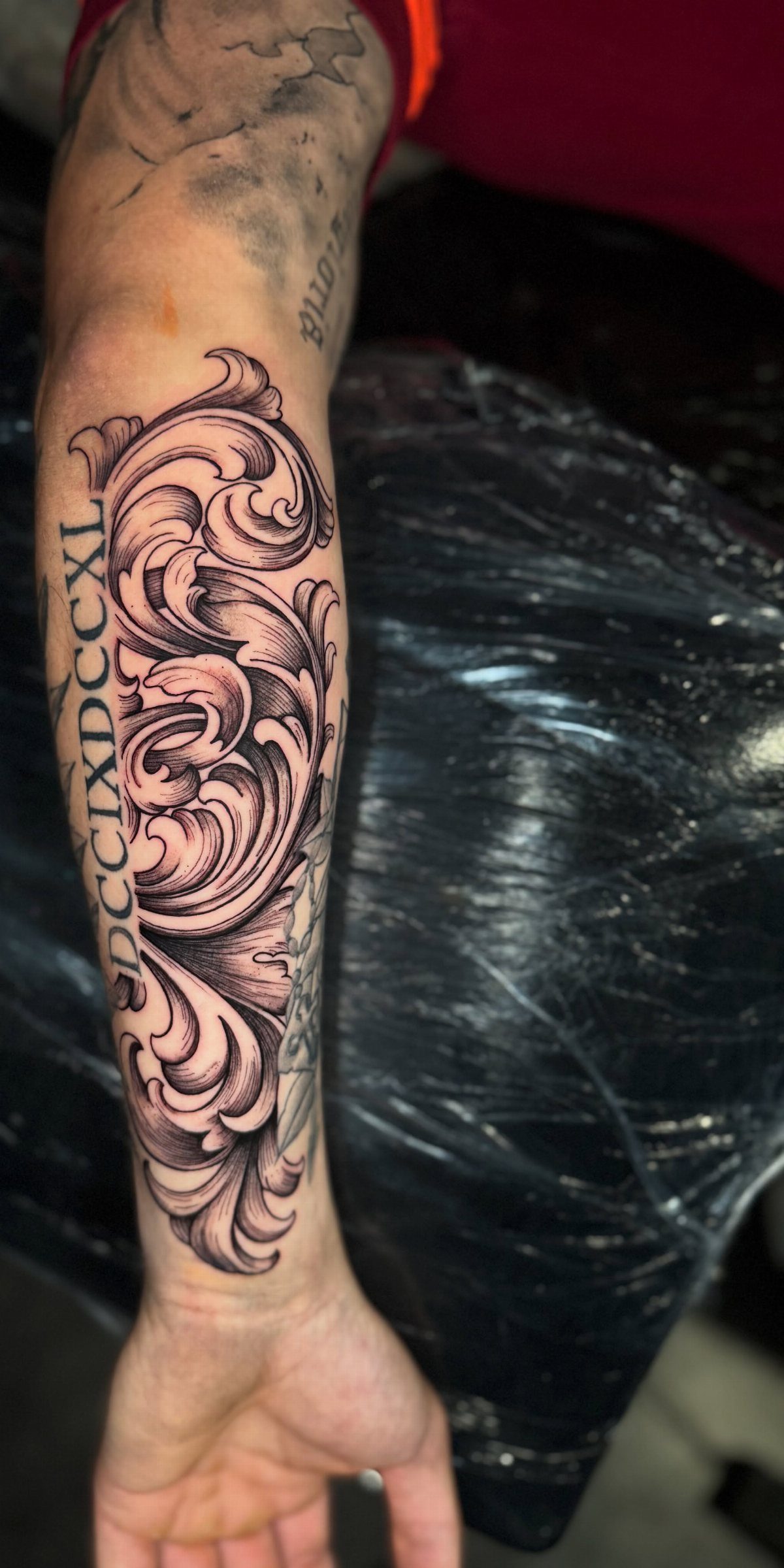 A man's forearm displaying a detailed black and grey filigree tattoo with Roman numerals.