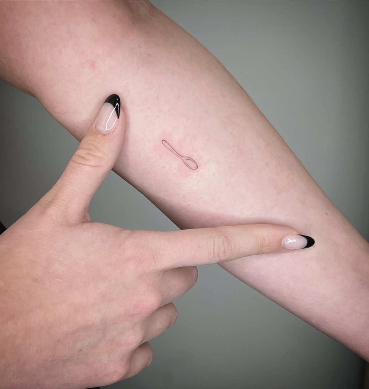 Sick Tattoo Art: 9 Styles That Redefine the Needle 1 A small minimalist spoon tattoo on a person's forearm being pointed at by two hands.