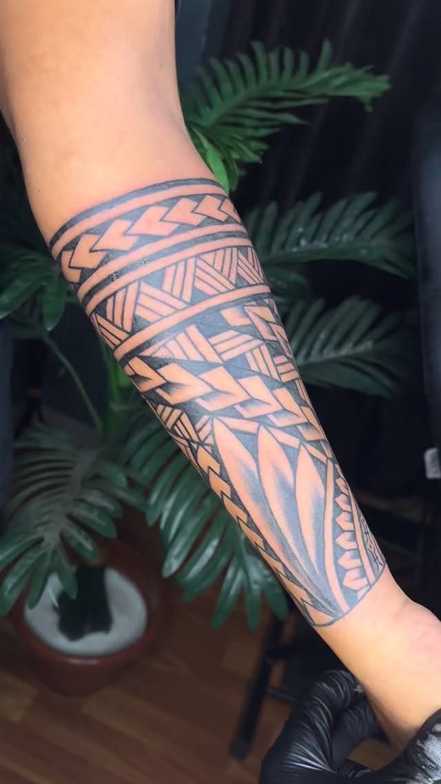 A forearm decorated with a large black geometric tribal pattern tattoo against green leaves.