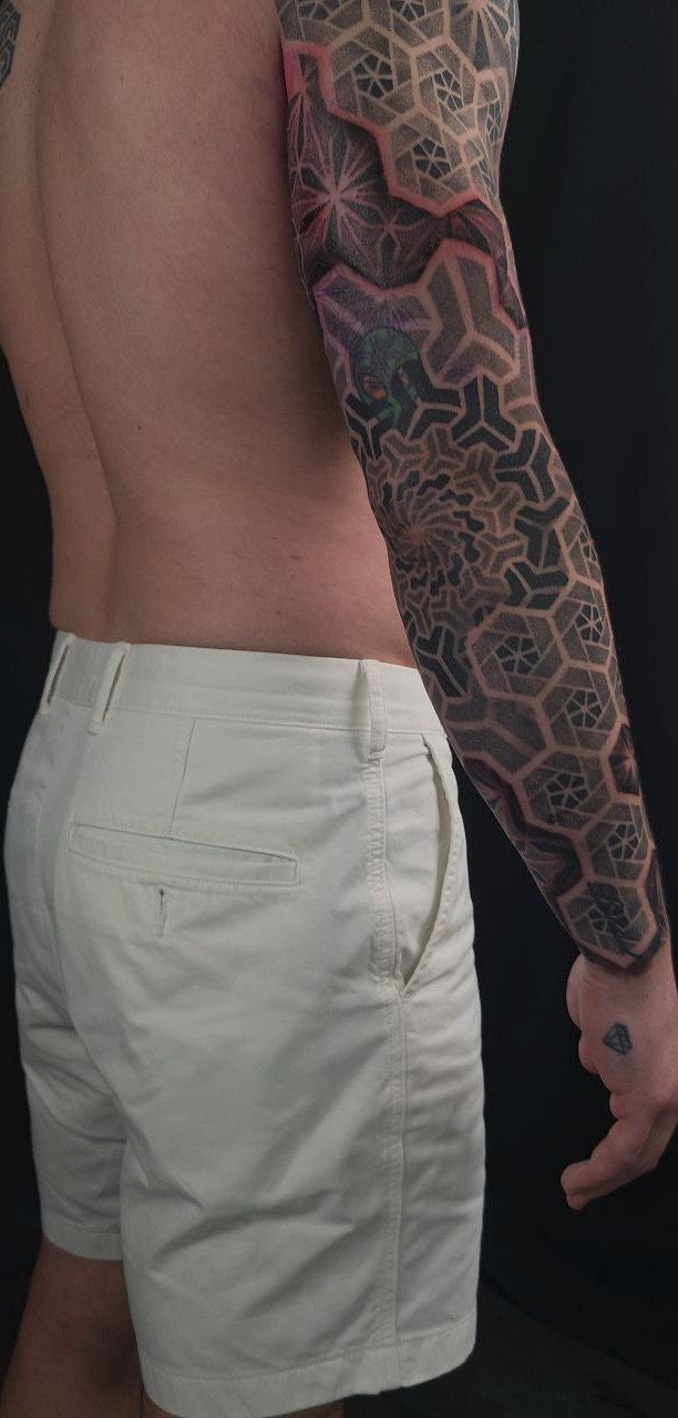 Man with a full sleeve geometric pattern tattoo on his arm standing against a black background.