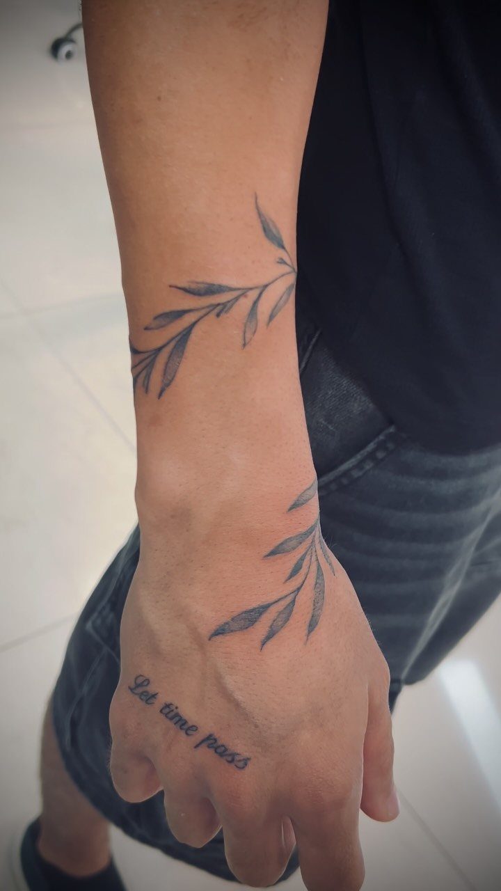 A person's wrist and hand featuring delicate leaf branch tattoos and text on the hand.