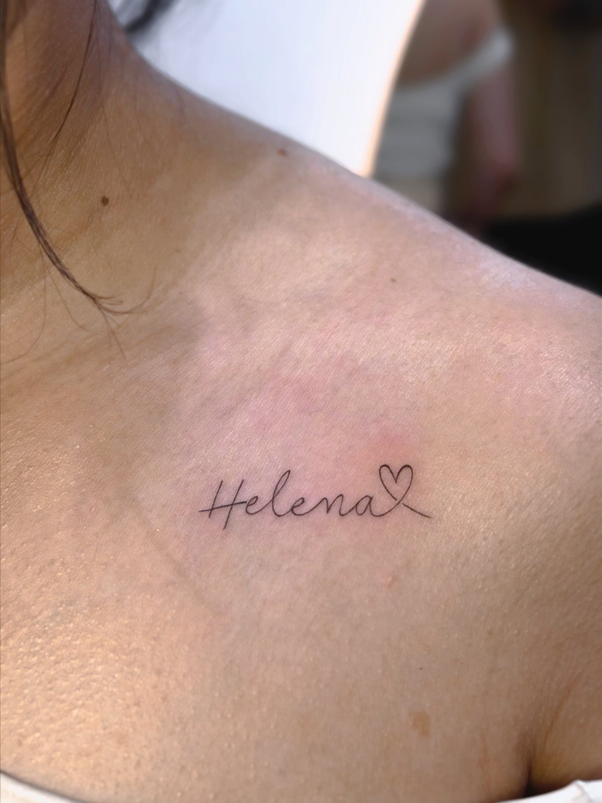 A delicate cursive tattoo of the name Helena with a small heart on a collarbone.