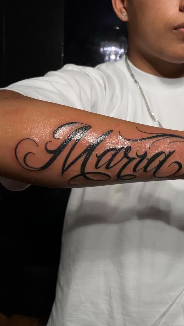 A large black cursive tattoo of the name Maria on a person's inner forearm.