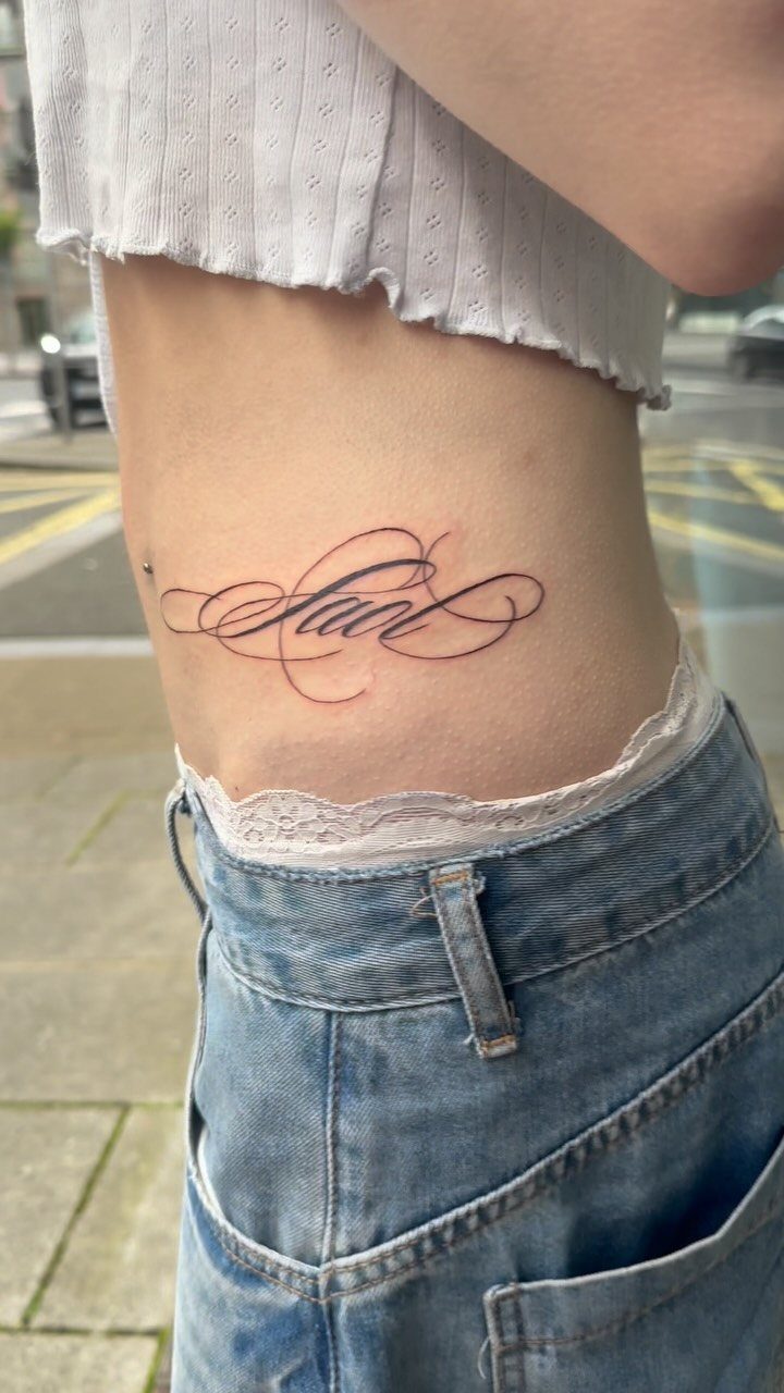 A cursive name tattoo on a person's side above blue denim jeans.