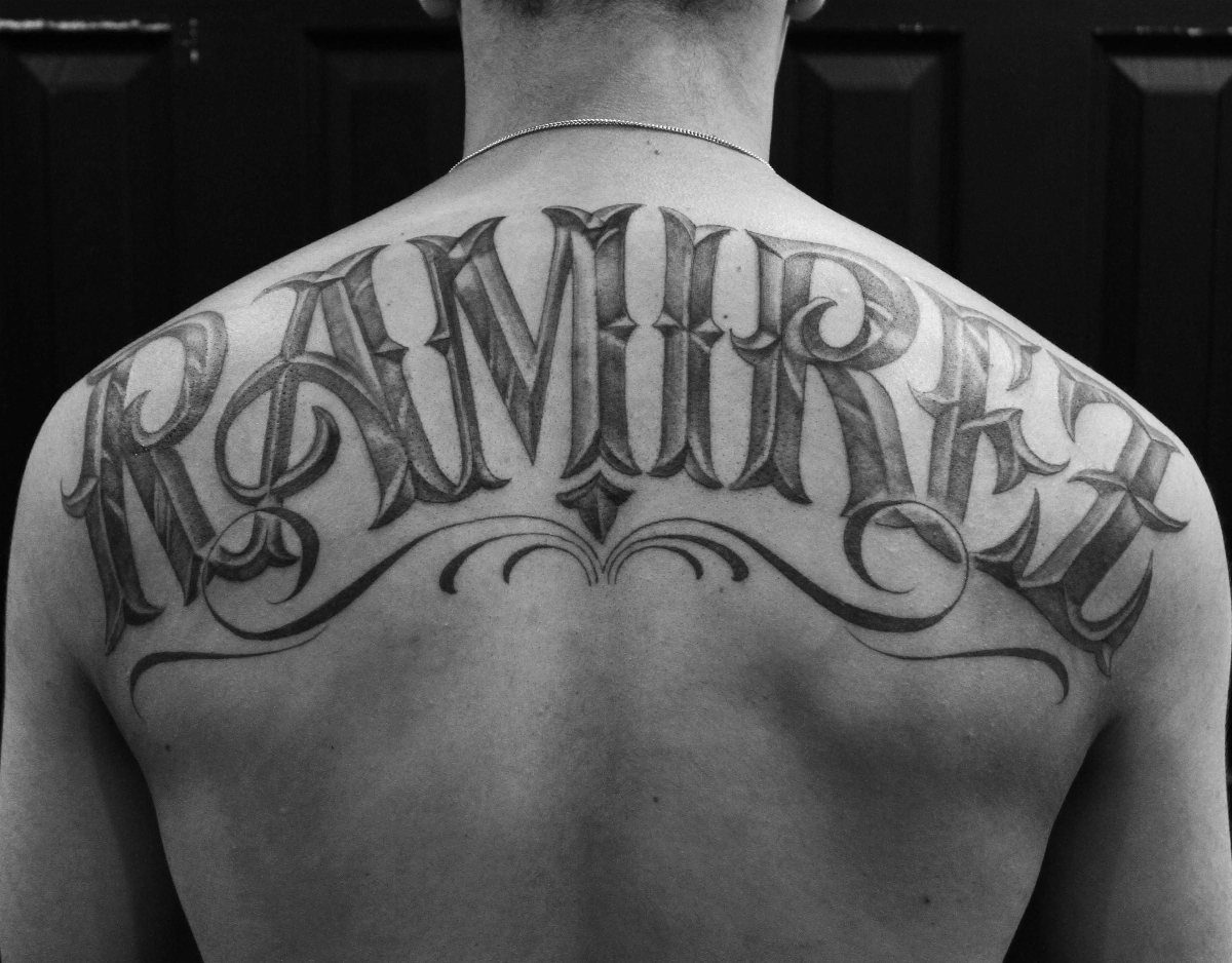 A large gothic style name tattoo reading RAMIREZ arched across a person's upper back.