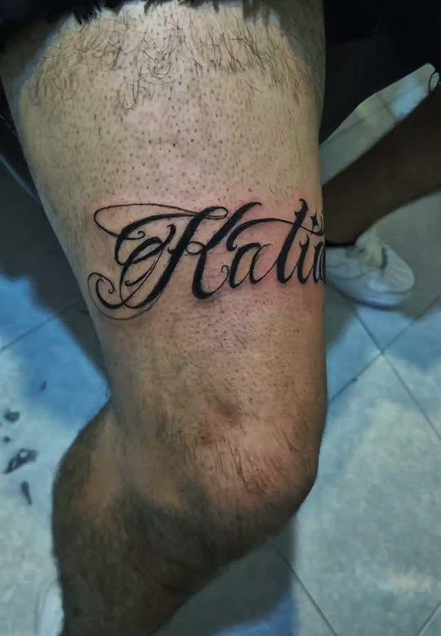 A large black cursive script tattoo reading Katia on a person's hairy thigh.