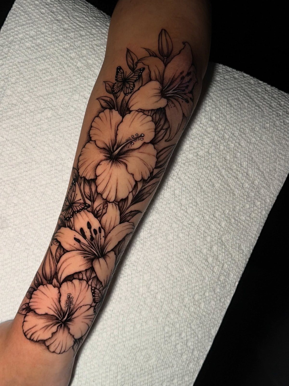 Tattoo Style Drawings: 6 Sketches Worth Keeping in Your Book 4 A detailed black ink floral tattoo with hibiscus and lilies on a person's forearm.