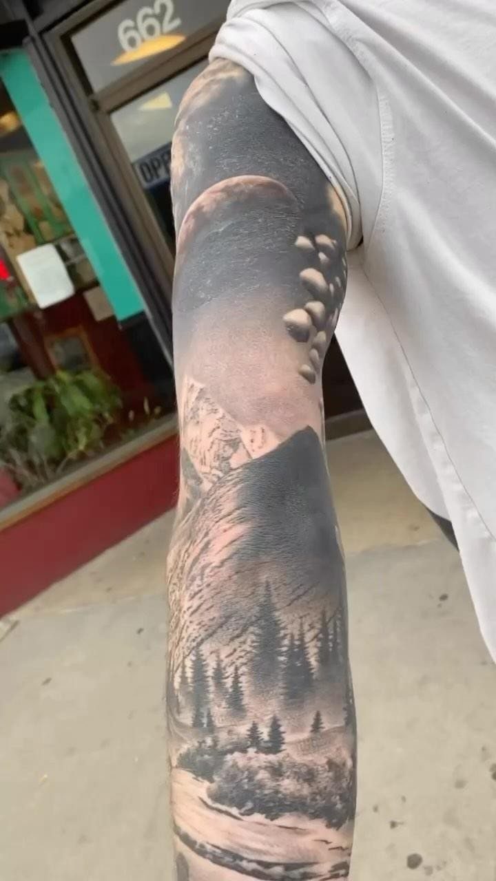 10 Tattoo Style Nature Ideas for Your Next Session 9 A detailed black and grey realism tattoo on an arm showing mountains, moon, and pine trees.