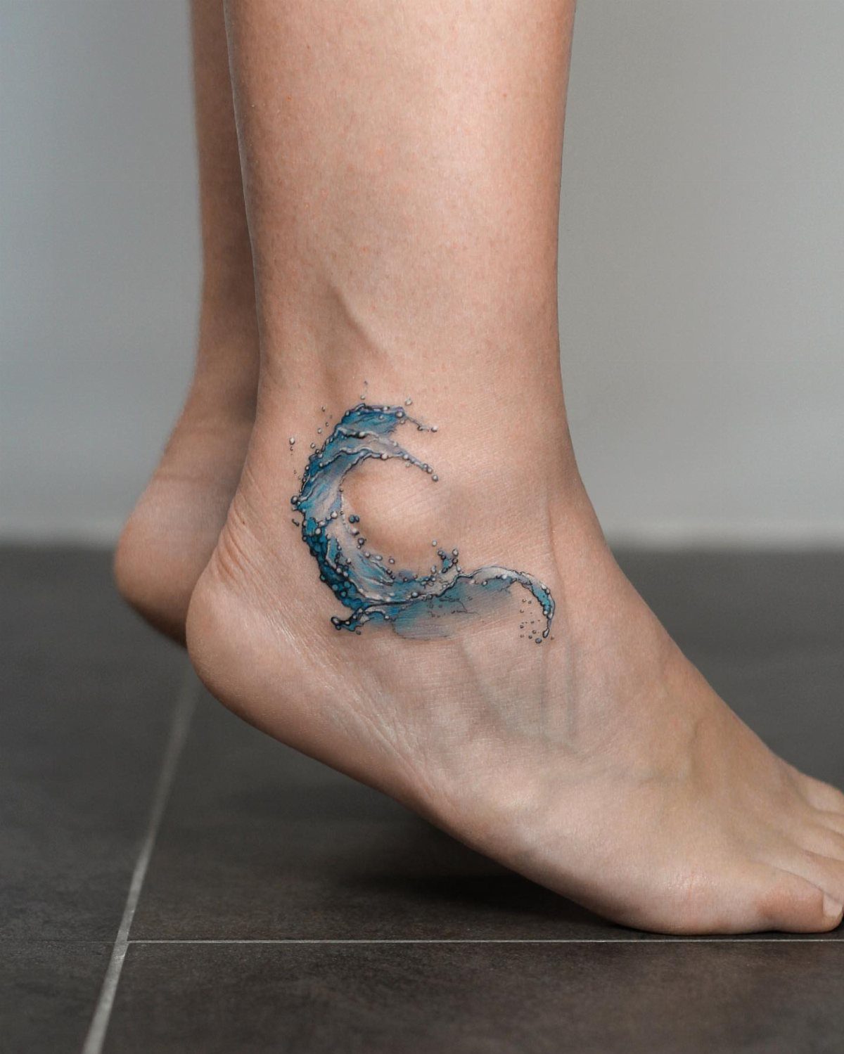 10 Tattoo Style Nature Ideas for Your Next Session 5 A blue watercolor wave tattoo on a person's ankle near the foot.