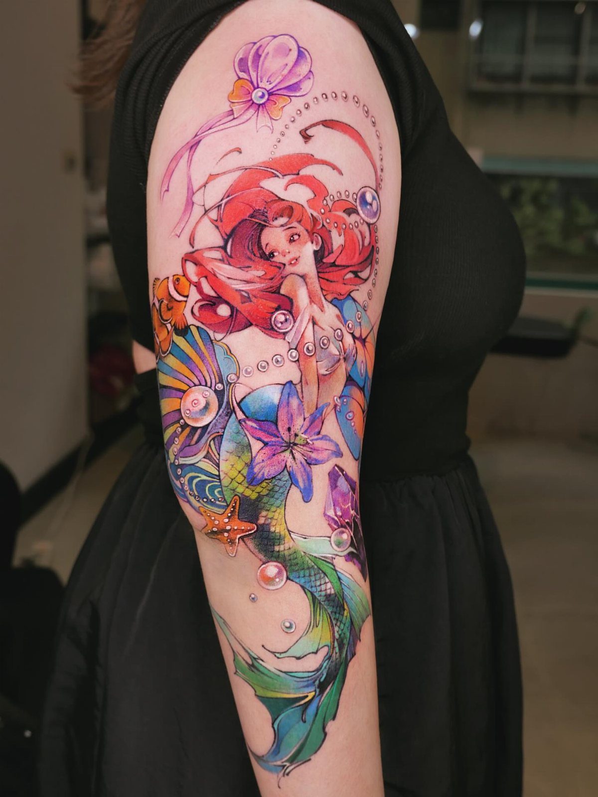 A colorful mermaid tattoo with red hair and seashells on a person's arm.