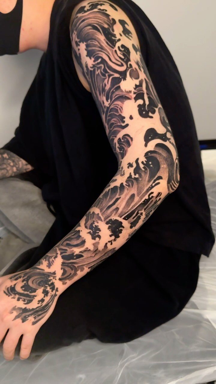 A person's arm featuring a large black and grey ocean wave sleeve tattoo.