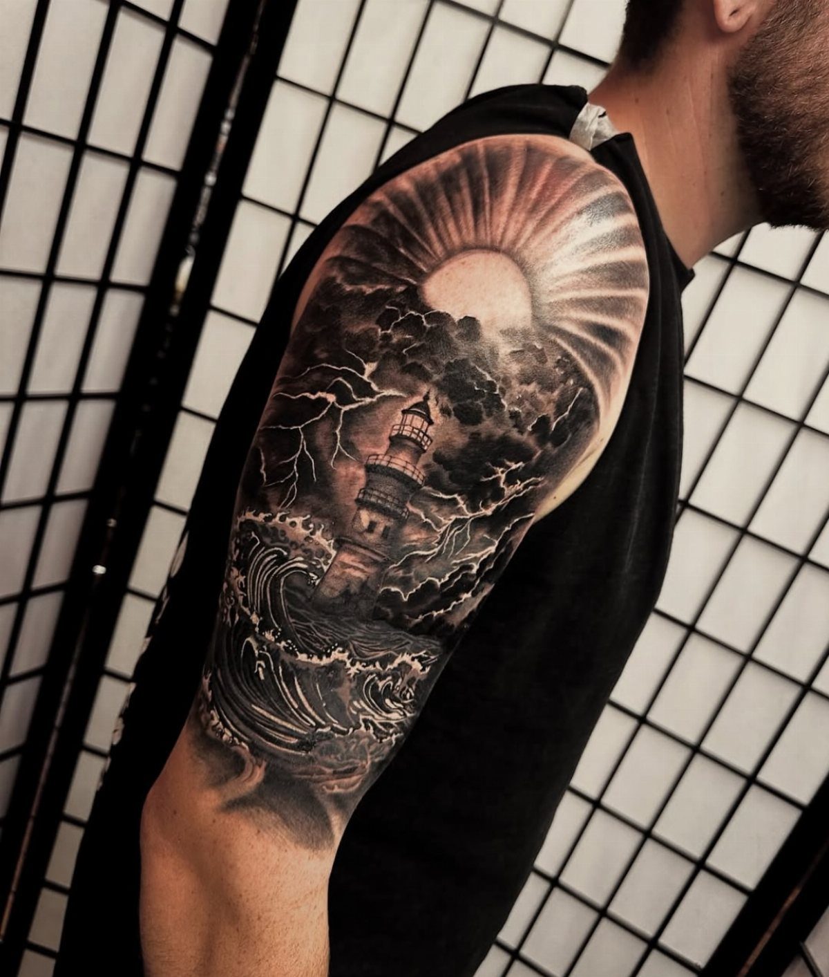 A detailed black and grey tattoo of a lighthouse in a stormy ocean with lightning.