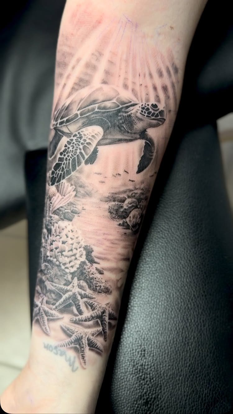 A realistic black and grey sea turtle and coral reef tattoo on a person's forearm.
