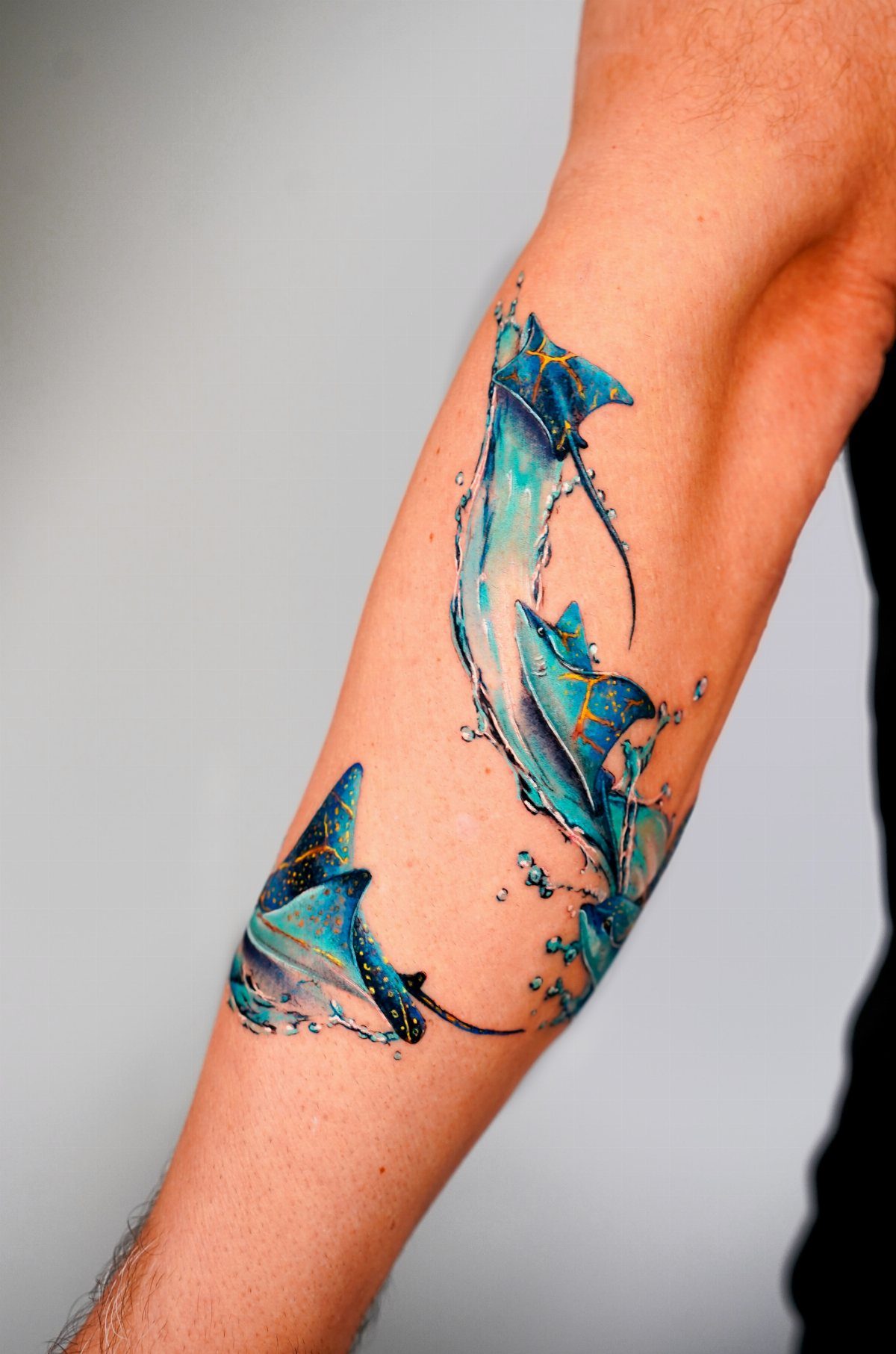 A vibrant blue and teal stingray tattoo with water splashes on a person's forearm.