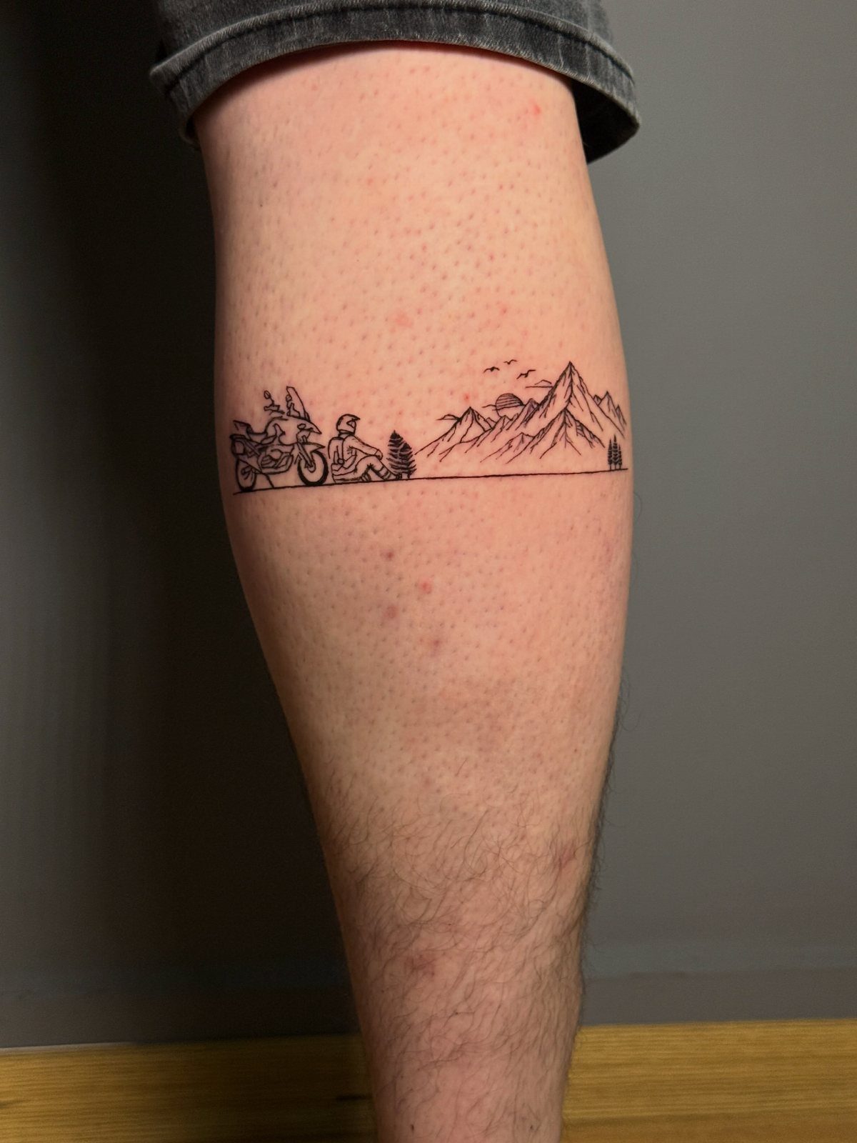 A black ink line art tattoo of a motorcycle and mountains on a person's leg.