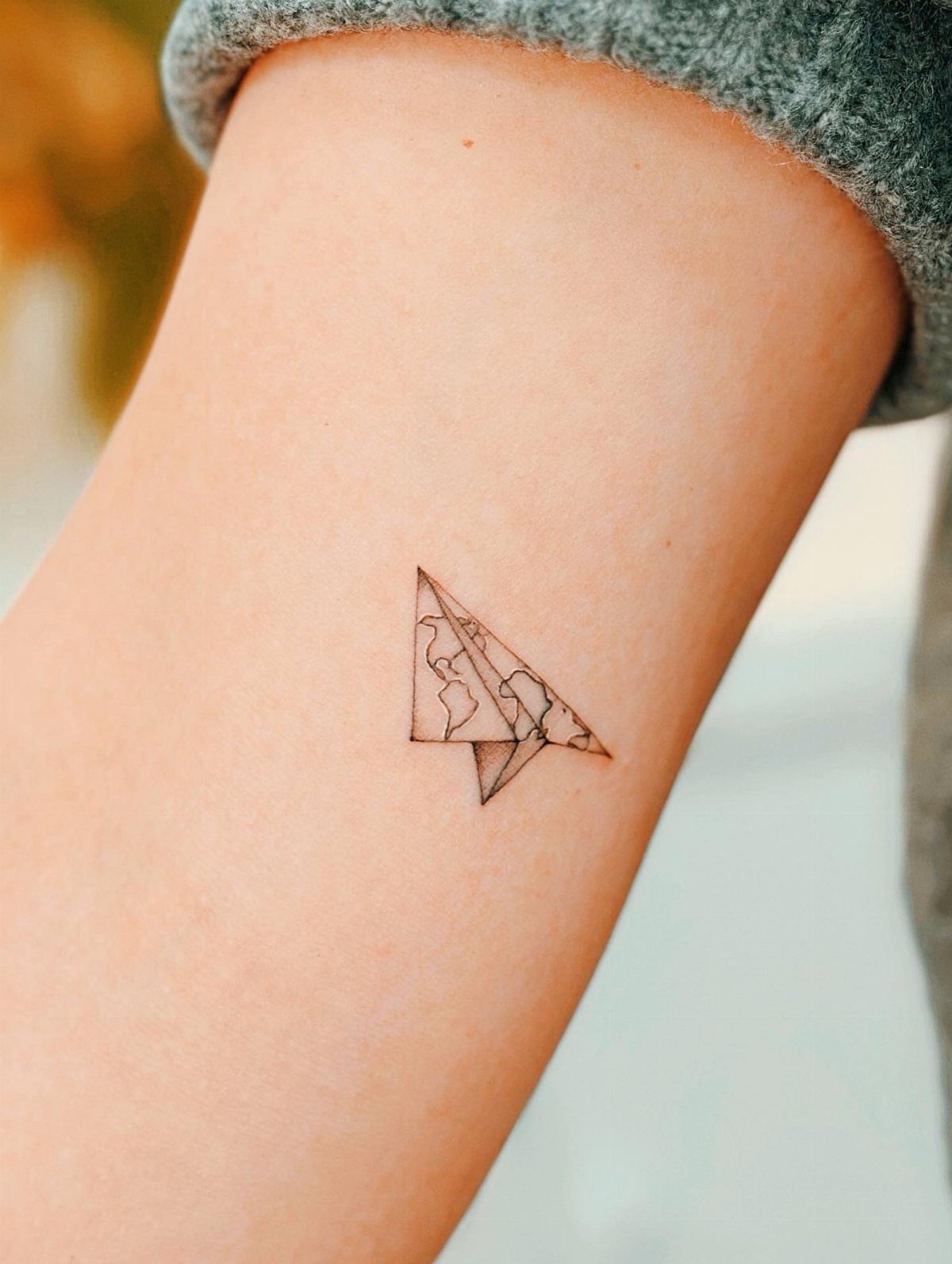 A minimalist fine-line tattoo of a paper plane with a map design on a person's arm.