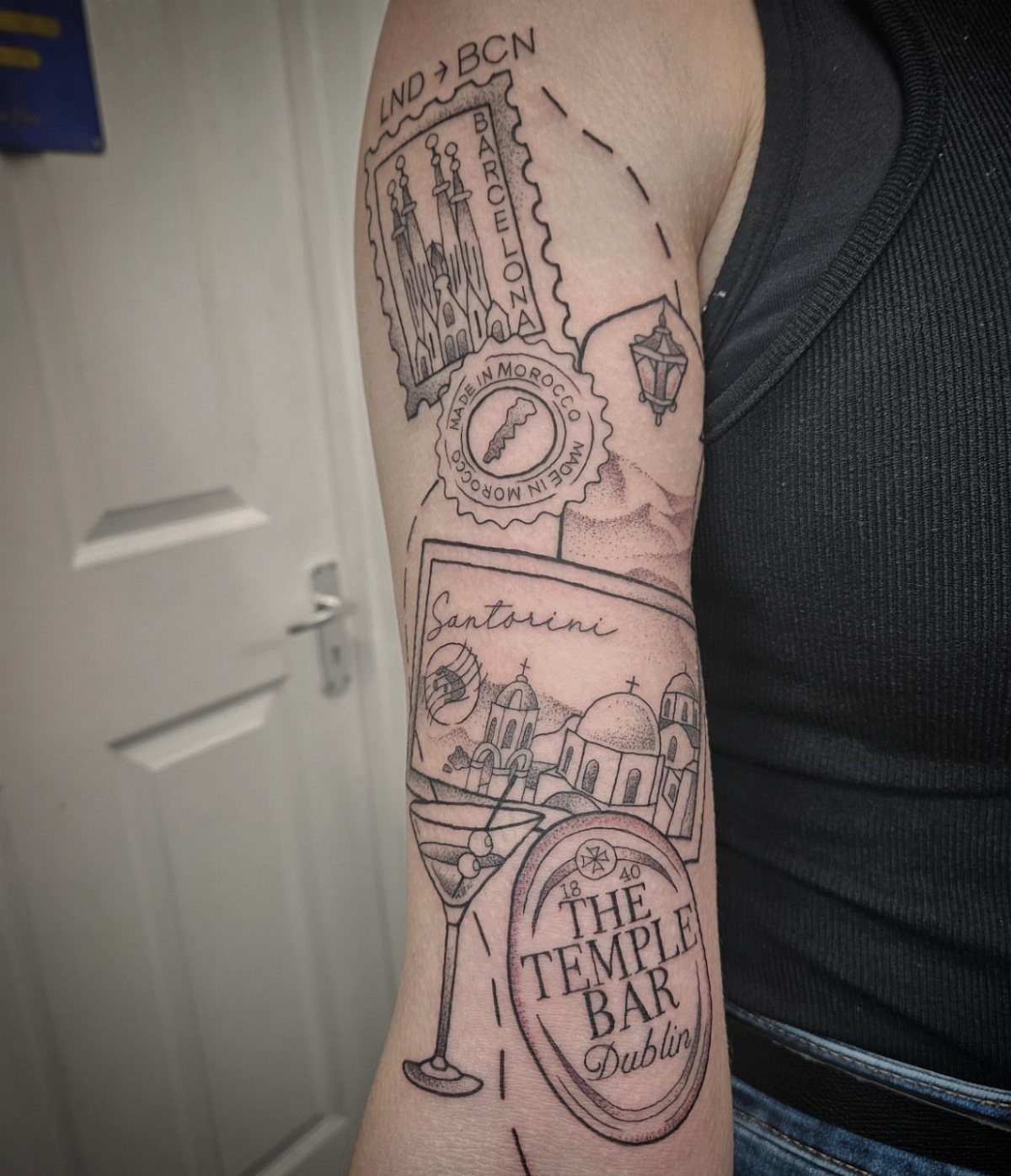 A detailed black ink sleeve tattoo on an arm featuring various travel and destination motifs.