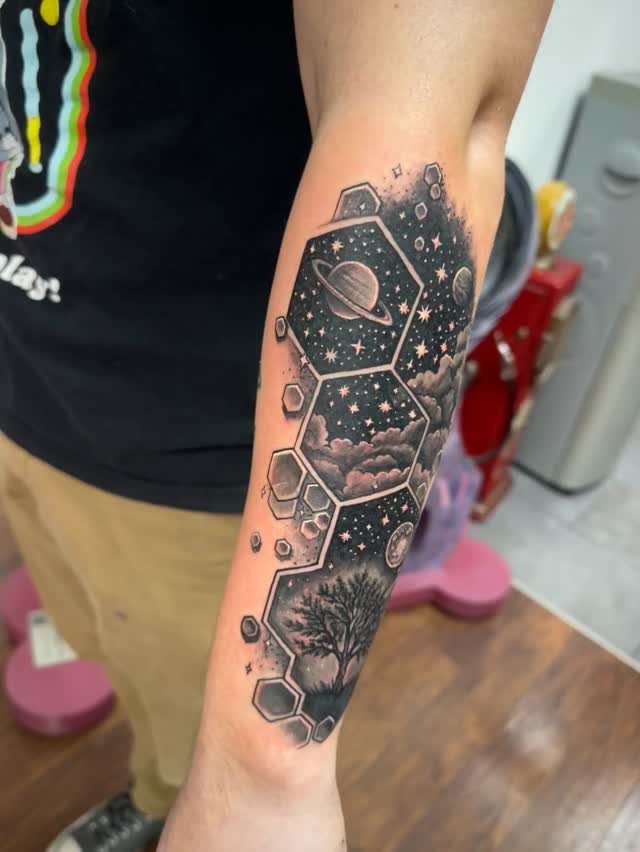 Universe Tattoo Pattern: 8 Space-Inspired Designs for Your Next Ink 5 A black and grey forearm tattoo depicting a cosmic nebula and planets inside hexagonal geometric shapes.