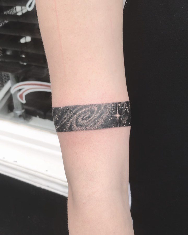 Universe Tattoo Pattern: 8 Space-Inspired Designs for Your Next Ink 3 A black ink band tattoo on an arm depicting a stippled spiral galaxy with stars.