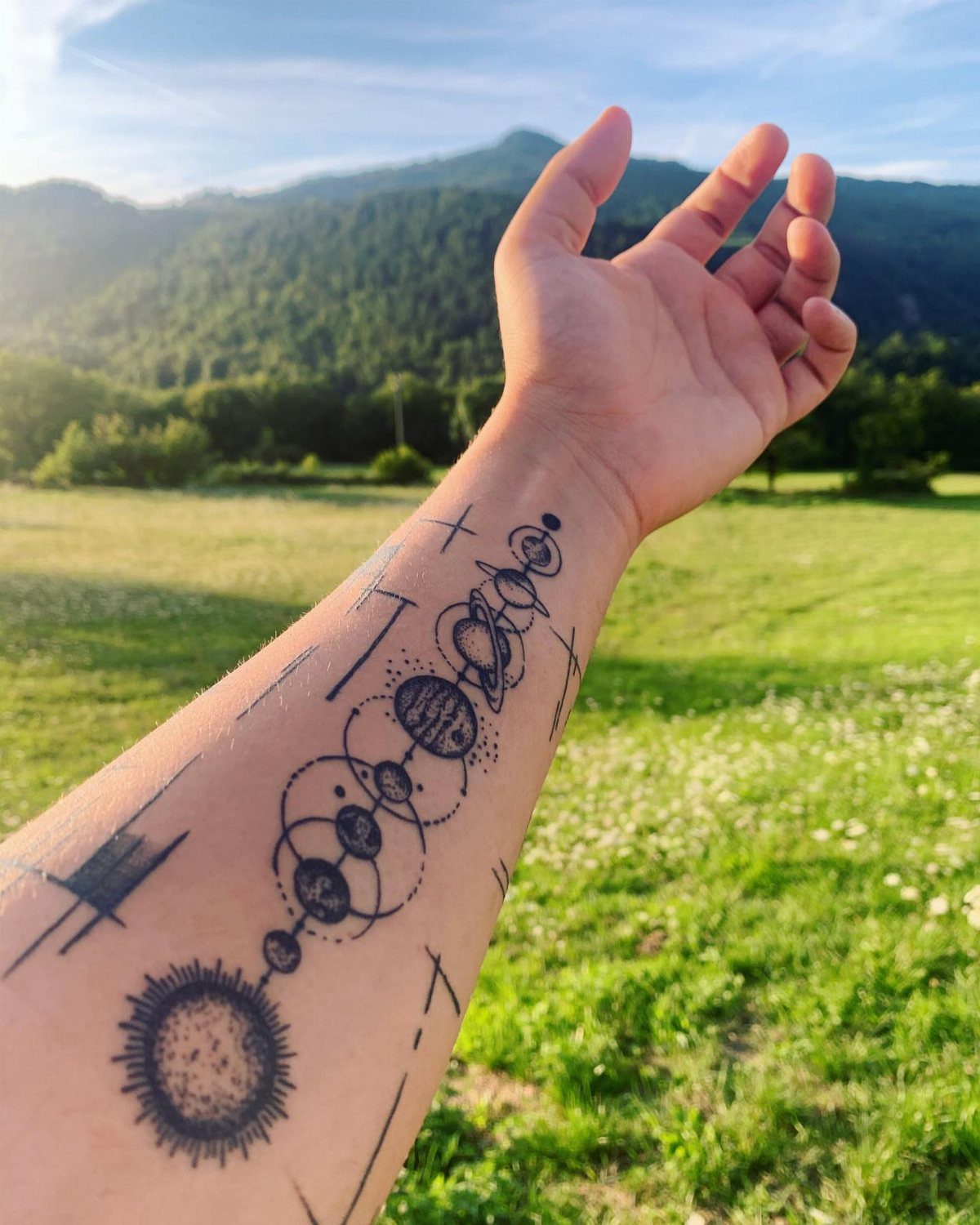 Universe Tattoo Pattern: 8 Space-Inspired Designs for Your Next Ink 6 A forearm tattoo featuring stippled planets and geometric lines against a natural outdoor landscape.