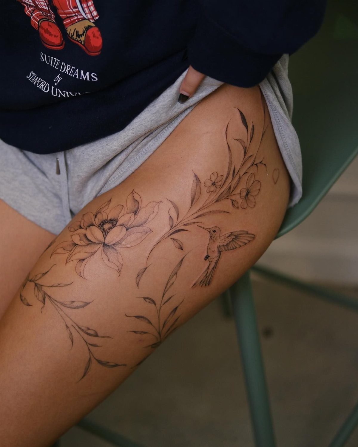 Black ink tattoo of a flower, hummingbird, and swirling vines on a person's thigh.