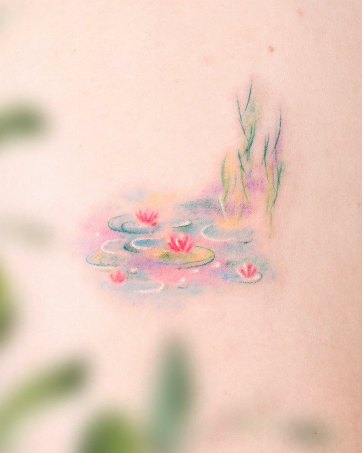 Watercolor pink water lily tattoo with green lily pads and grass on skin.