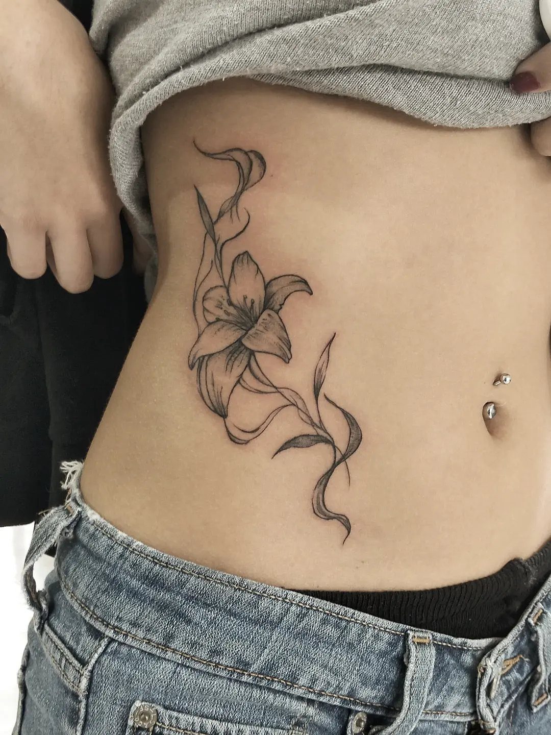 5 Wrap Around Waist Tattoo Ideas That Follow Your Body's Curve 1 A fine line black ink lily tattoo with flowing stems on a person's abdomen.