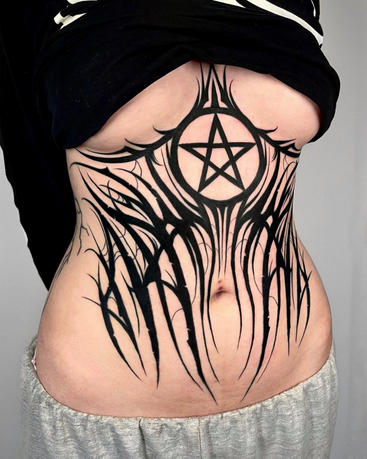 5 Wrap Around Waist Tattoo Ideas That Follow Your Body's Curve 3 A large black tribal tattoo with a pentagram design covering a person's abdomen and waist.